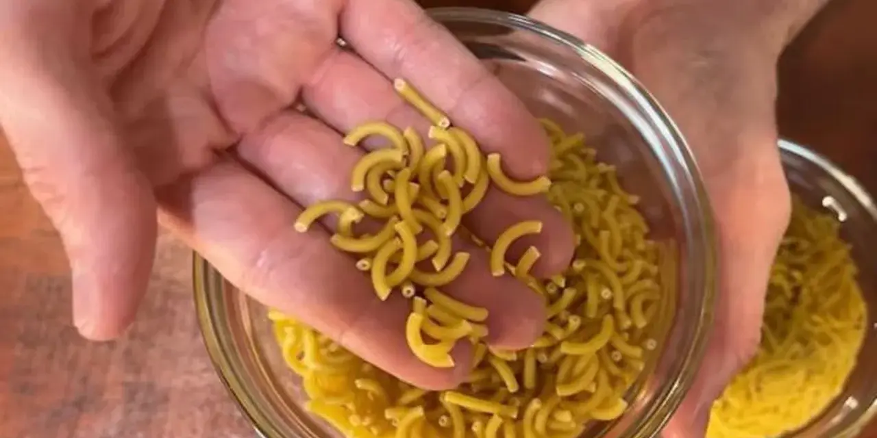 Mano sosteniendo fideos cortos y curvos sobre un bol de fideos similares.