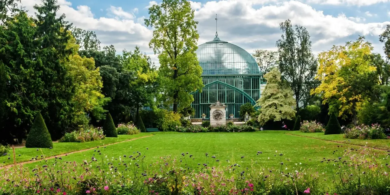 Un jardín exuberante en el Bois de Boulogne, con una gran estructura de cristal verde y un cielo azul con nubes.