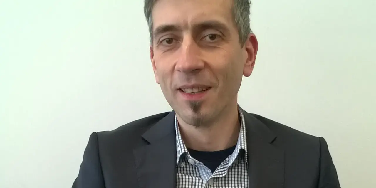 Maciej Kasprzak w czarnym garniturze i koszuli w kratę, z charakterystyczną bródką.