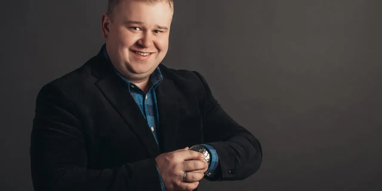 Uśmiechnięty mężczyzna w czarnym garniturze i niebieskiej koszuli, sprawdzający zegarek. Podpowiadamy, jak dobrać garnitur dla pana z brzuszkiem.