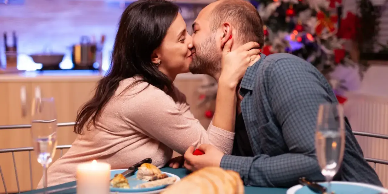 Romantyczny wieczór, kolacja przy świecach i choince. Para celebruje dzień pocałunku, mężczyzna trzyma w dłoni czerwone pudełeczko.