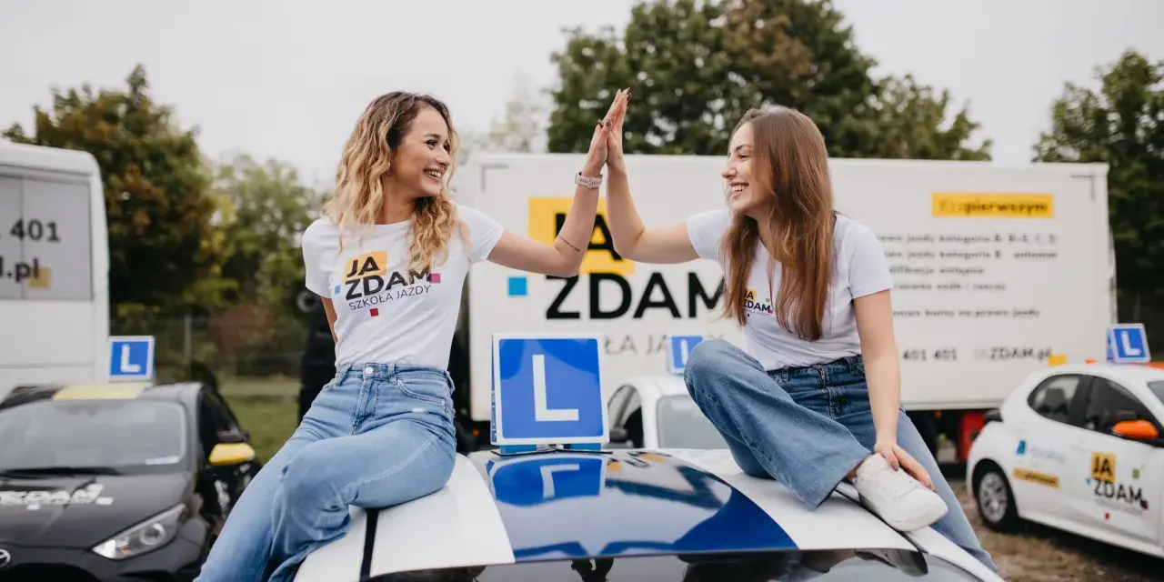 Dwie uśmiechnięte dziewczyny świętują sukces na dachu samochodu szkoleniowego. W tle ciężarówka z informacją o kursach na różne kategorie prawa jazdy.