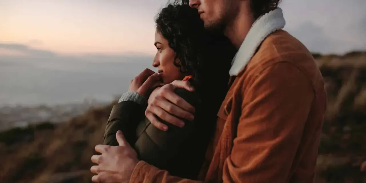 A couple embraces, looking out at a hazy landscape. This image shows how to feel more connected through shared moments and physical closeness.