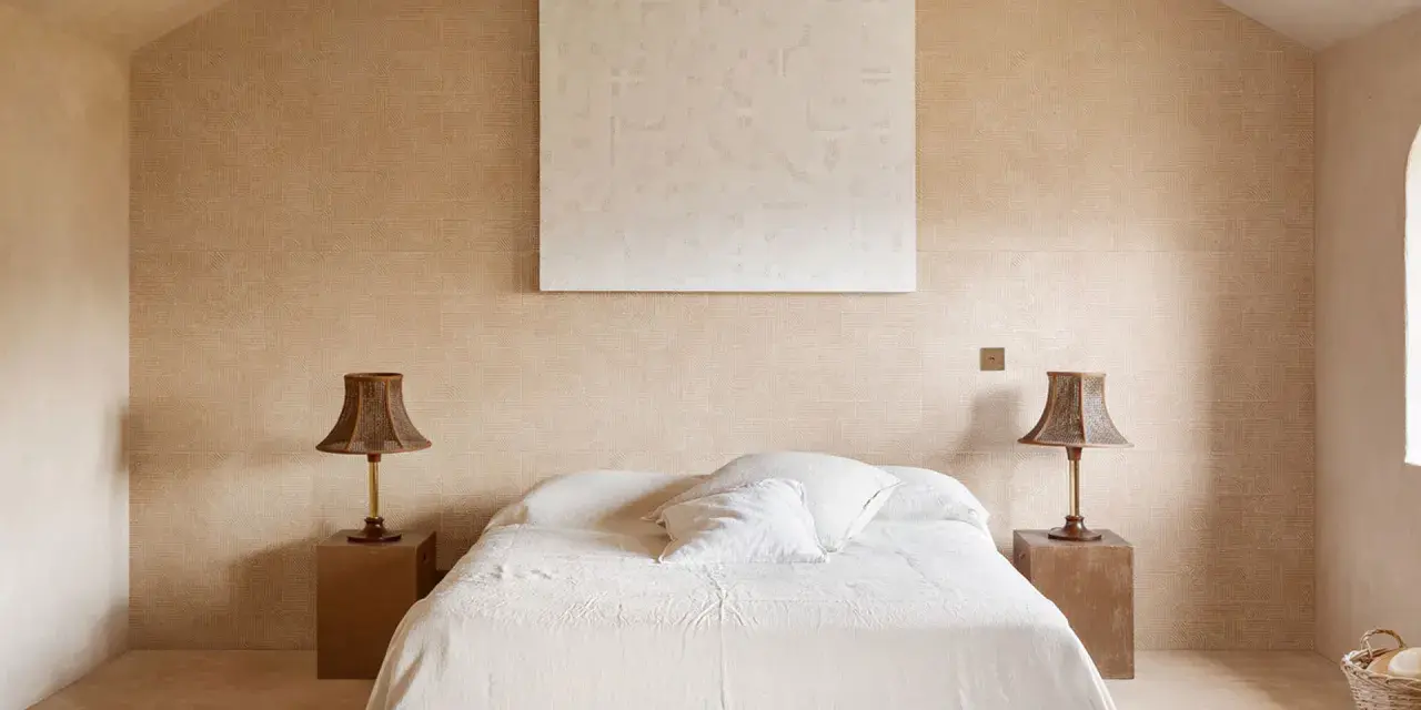 Camera da letto aesthetic con pareti color sabbia, letto bianco e due lampade in vimini. Un quadro astratto completa l'arredo.