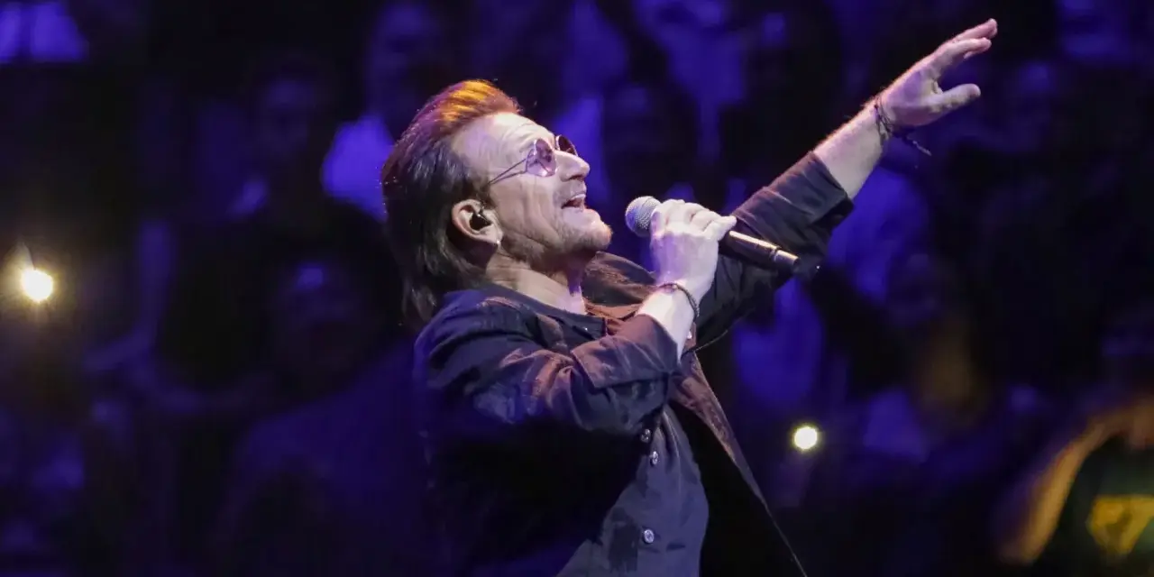 Bono z U2 śpiewa na koncercie, zapowiadając u2 nowa płyta. Tłum w ciemnościach reaguje na jego występ.