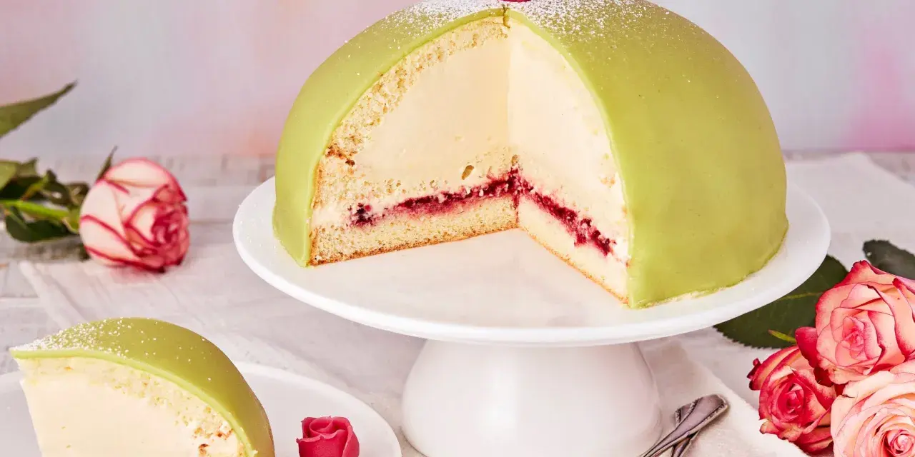 Ein Anschnitt eines grünen Prinzessinnenkuchens mit Sahnefüllung und Himbeermarmelade, verziert mit einer Zuckerrose. Ein Stück liegt auf einem Teller daneben.