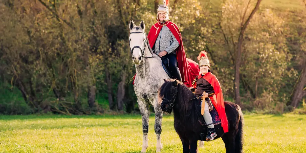Zwei Reiter in römischer Kleidung, ein Erwachsener auf einem Schimmel und ein Kind auf einem Pony, reiten auf einer sonnigen Wiese. Ein fröhlicher Anblick, wie bei einem Martin Fest.