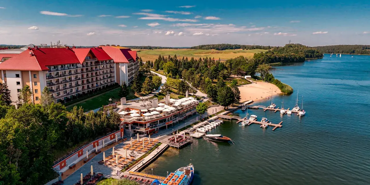 Hotel na wyspie Mikołajki z czerwonym dachem, otoczony zielenią, z mariną pełną łodzi i statkiem wycieczkowym na jeziorze.