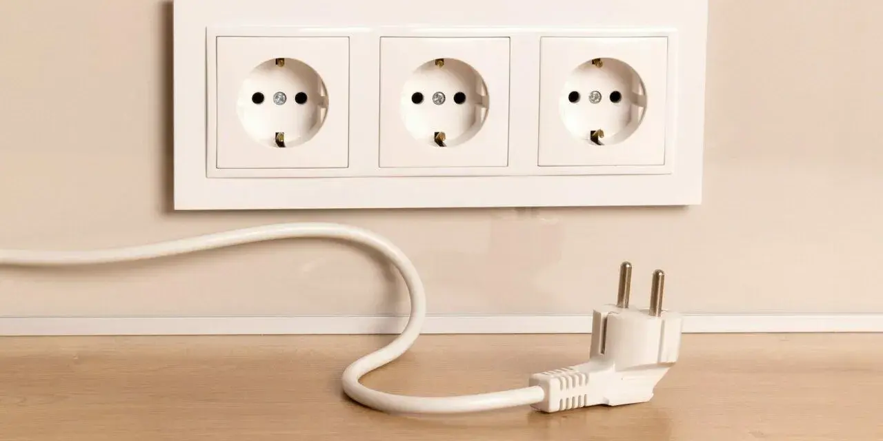 Biały kabel z wtyczką leży na drewnianym blacie obok potrójnego gniazdka. Jak schować kable, by wyglądały estetycznie?