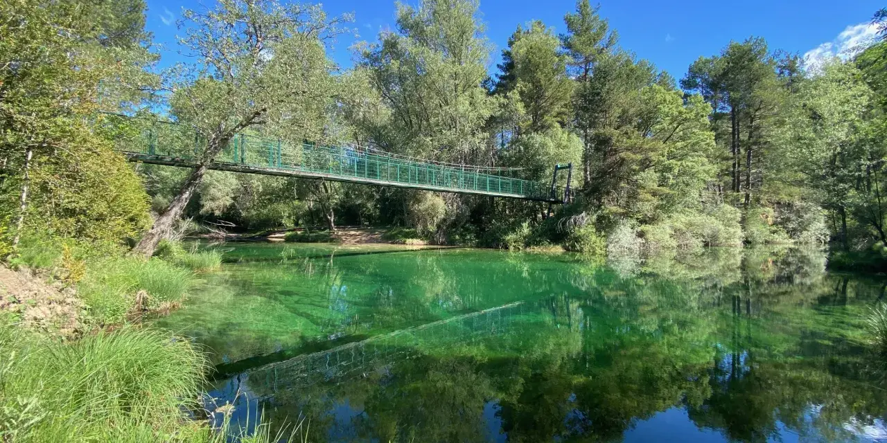 Puente verde cruza un río de aguas cristalinas y verdes, reflejando el bosque exuberante. Un lugar idílico para explorar el **alto tajo**.