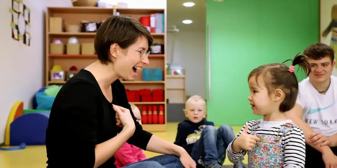 Eine Frau lacht und gestikuliert, während sie mit einem Kleinkind sprechen übt. Ein Junge und ein Mann sitzen im Hintergrund.