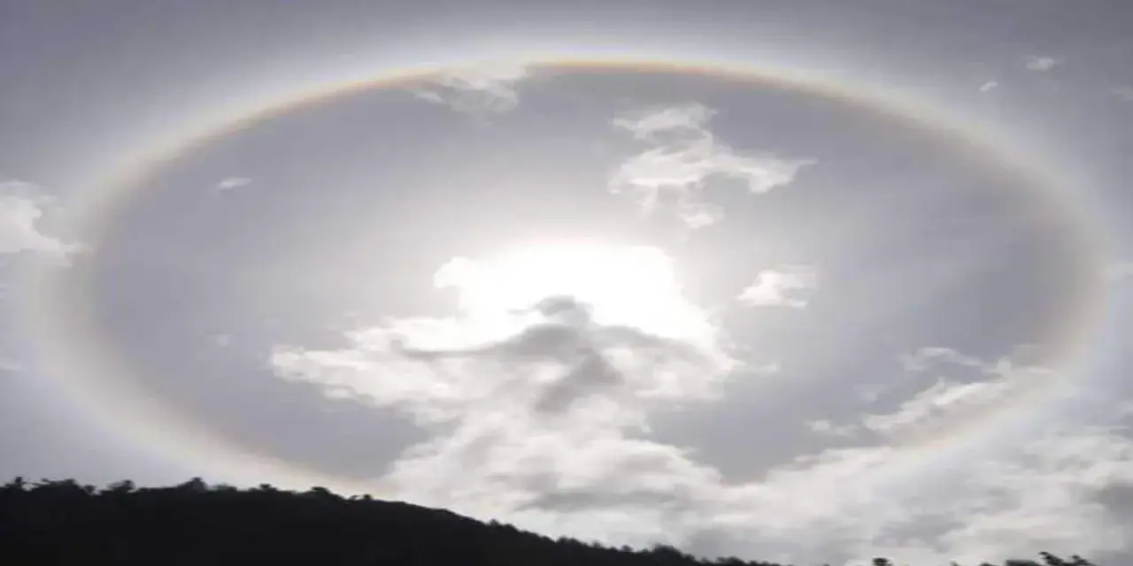Un alone arcobaleno circonda il sole tra le nuvole, creando un effetto magico sopra una collina scura.