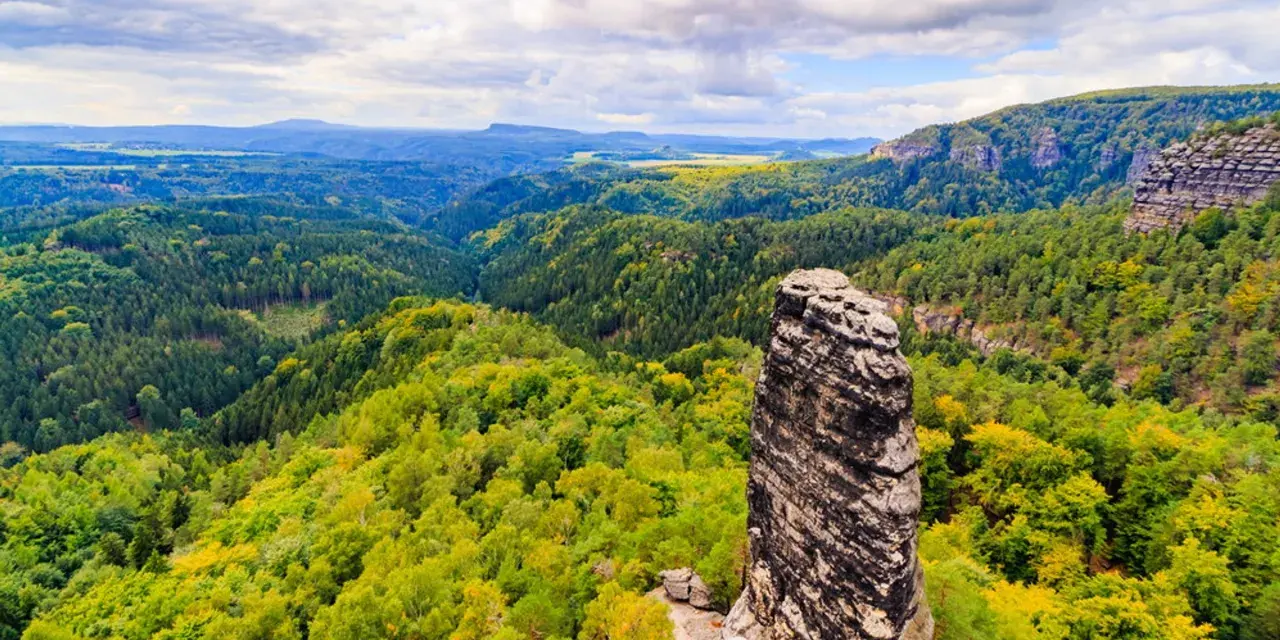 Skalny filar góruje nad zielonymi lasami w Czechach, blisko granicy. Krajobraz pełen jest drzew i skalistych zboczy.