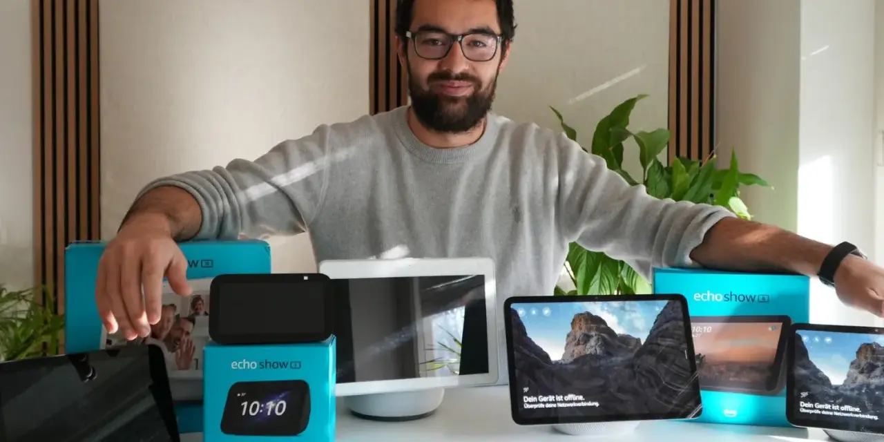 Mann präsentiert verschiedene Echo Show Generationen. Mehrere Geräte und Verpackungen sind auf dem Tisch zu sehen.
