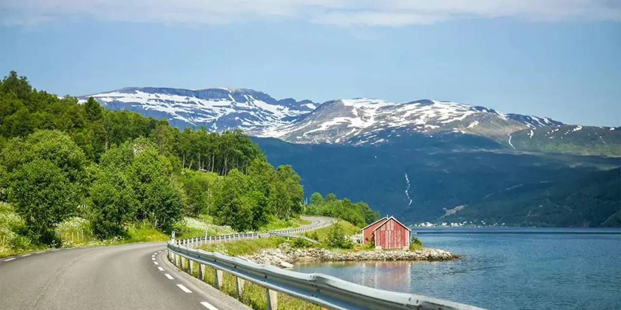 En slingrande väg längs en fjord i Norge, med snöklädda berg i bakgrunden. En röd stuga vid vattnet. Perfekt för en roadtrip norge rutt.
