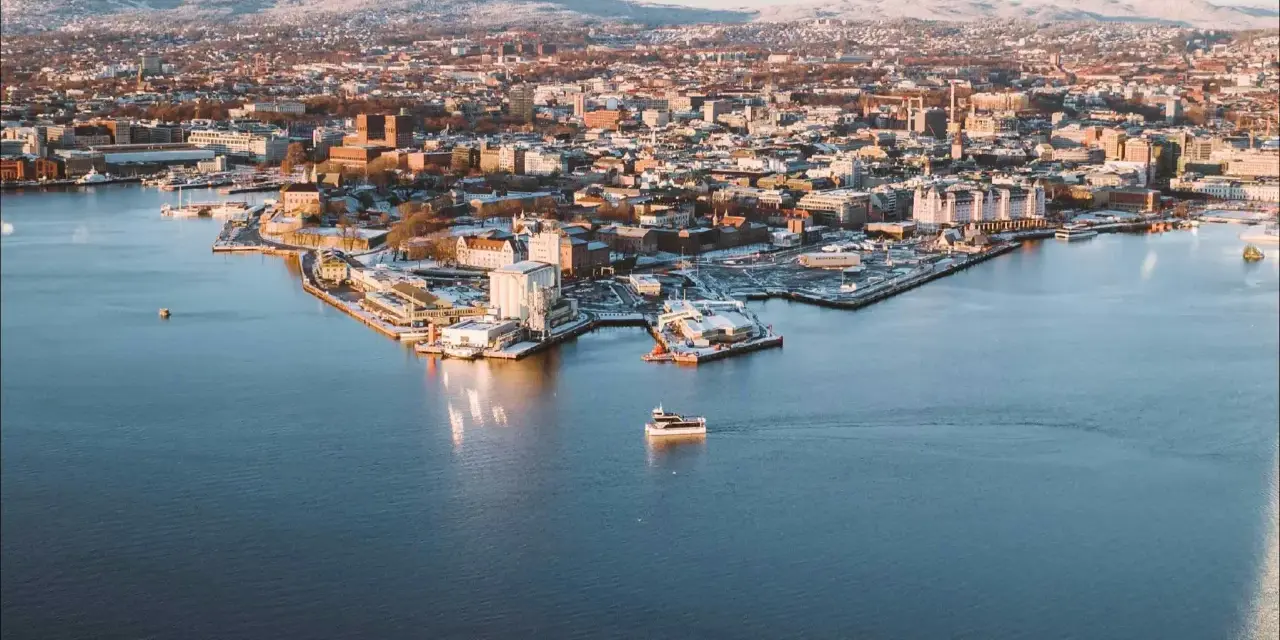Widok na zimowe Oslo z wody. Mała łódka płynie po spokojnym akwenie, w tle rozciąga się miasto i ośnieżone wzgórza.