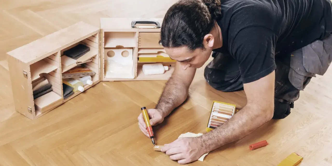 Mann repariert Parkett mit einem Reparaturstift und einer Farbauswahl. Werkzeugkasten mit Zubehör daneben.