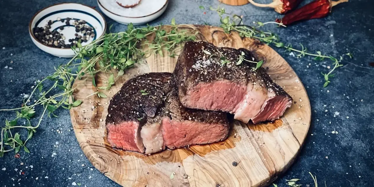 Zwei saftige Rib-Eye-Steaks, perfekt gegrillt, liegen auf einem Holzbrett. Mit Meersalz und Pfeffer bestreut, garniert mit frischem Thymian.