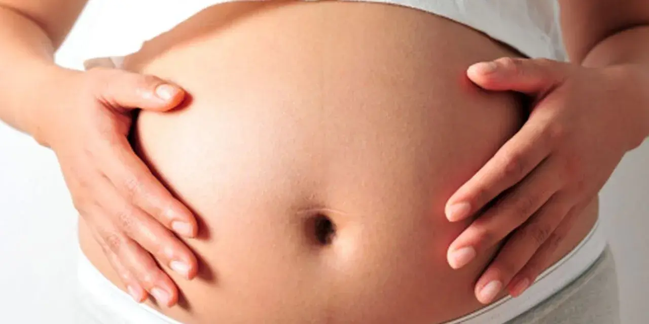 Brzuch ciężarnej kobiety, dłonie delikatnie go obejmują. Warto wiedzieć, jak rozpoznać sączące się wody płodowe.