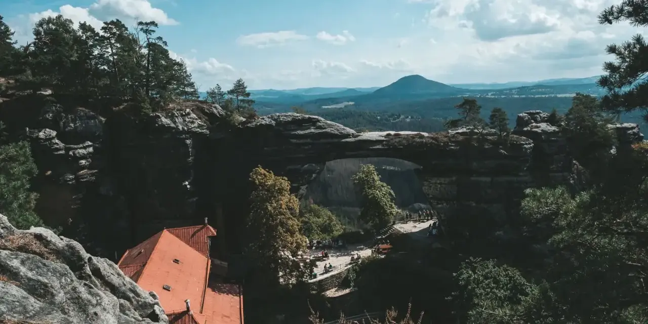 Majestatyczna kamienna brama w Czechach, otoczona zielenią i górami, z widokiem na urokliwy domek z czerwonym dachem.