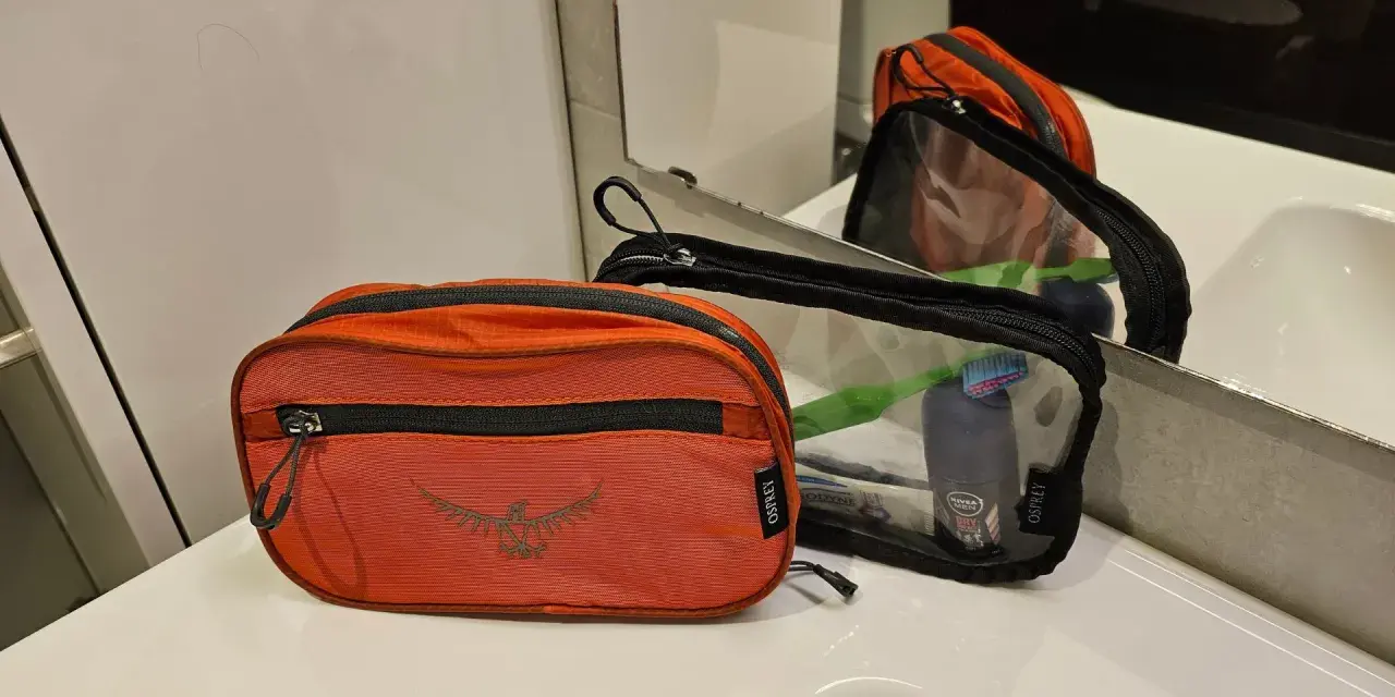 Två Osprey-necessärer, en orange och en genomskinlig, ligger på en vit bänkskiva. Den genomskinliga necessären innehåller tandborste och deodorant.