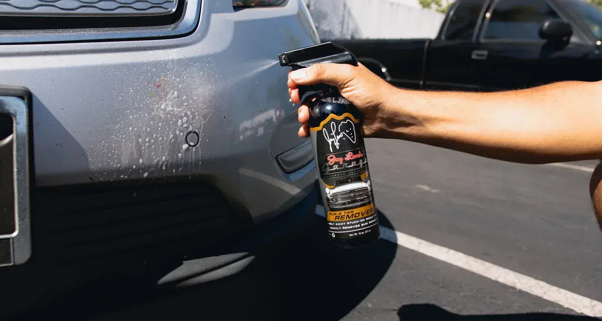 A hand sprays bug remover onto a car bumper, tackling stubborn bugs from a road trip.