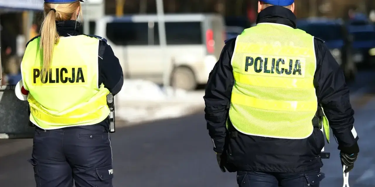 Dwóch policjantów w kamizelkach odblaskowych z napisem "POLICJA" stoi na drodze.