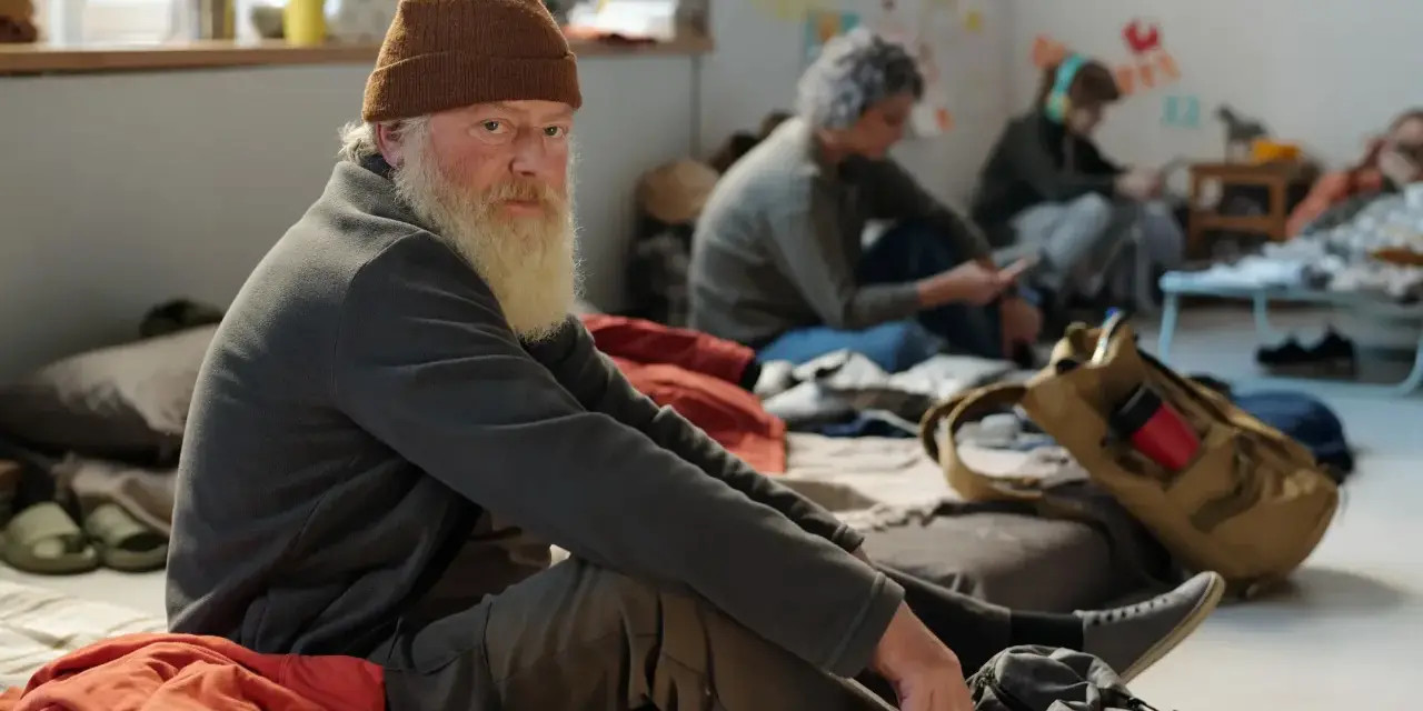 Älterer Mann mit Bart und Mütze sitzt auf dem Boden, umgeben von anderen Menschen. Eine Szene, die die Frage aufwirft: Wohin ohne Geld und Wohnung?
