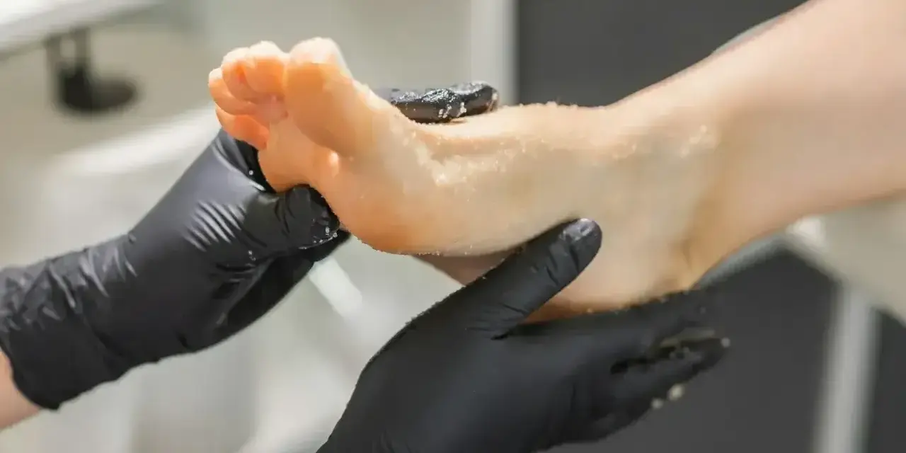 Dłonie w czarnych rękawiczkach wykonują peeling stopy. To właśnie jest pedicure co to znaczy pielęgnacja stóp.