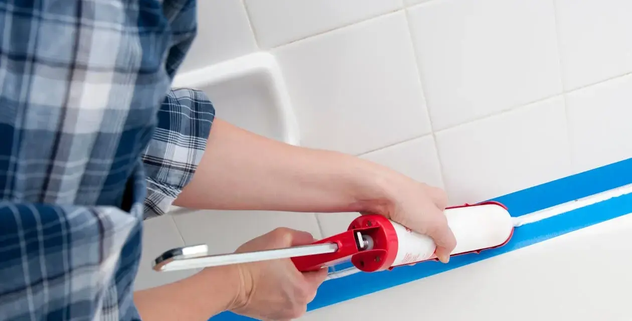 Osoba w kratę zakłada silikon w wannie, używając pistoletu. Błękitna taśma malarska wyznacza linię.