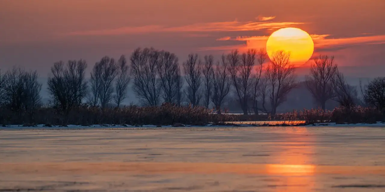 Zachód słońca nad zamarzniętym jeziorem. Piękna fotografia krajobrazowa z pomarańczowym niebem i sylwetkami drzew.