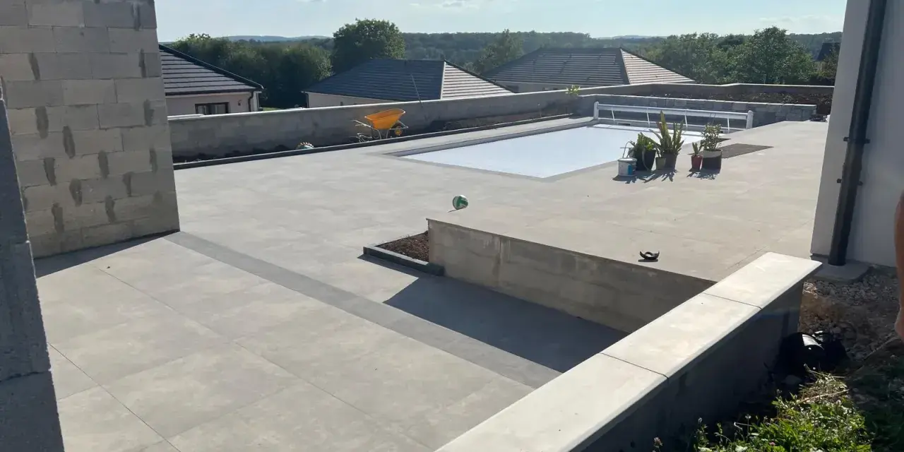 Vaste terrasse en béton gris avec piscine couverte, entourée de végétation et de maisons. Un chantier en cours.