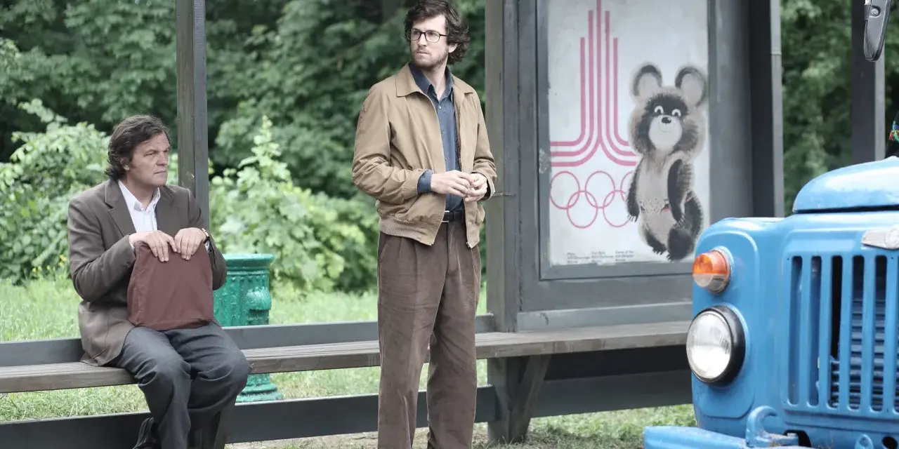Deux hommes attendent à un arrêt de bus, un panneau des JO de Moscou 1980 avec Misha l'ours en arrière-plan. L'affaire Farewell se déroule.
