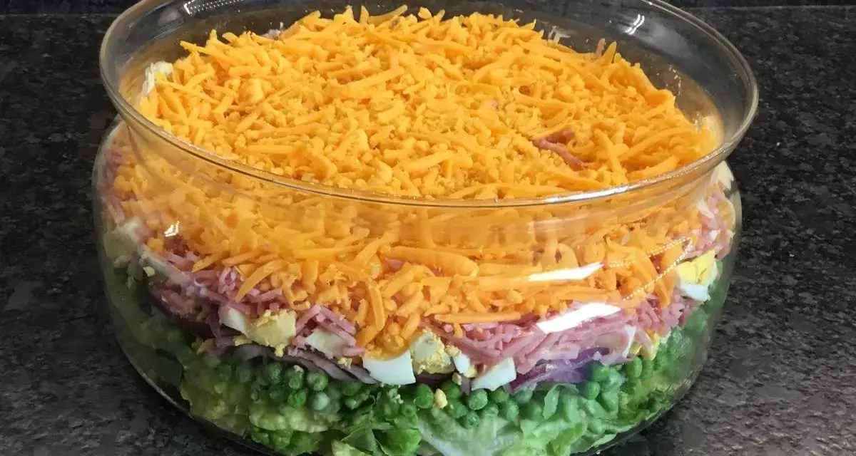Une salade 7 étages colorée dans un grand bol en verre, avec des couches de laitue, petits pois, oignons rouges, jambon, œufs durs et fromage râpé.