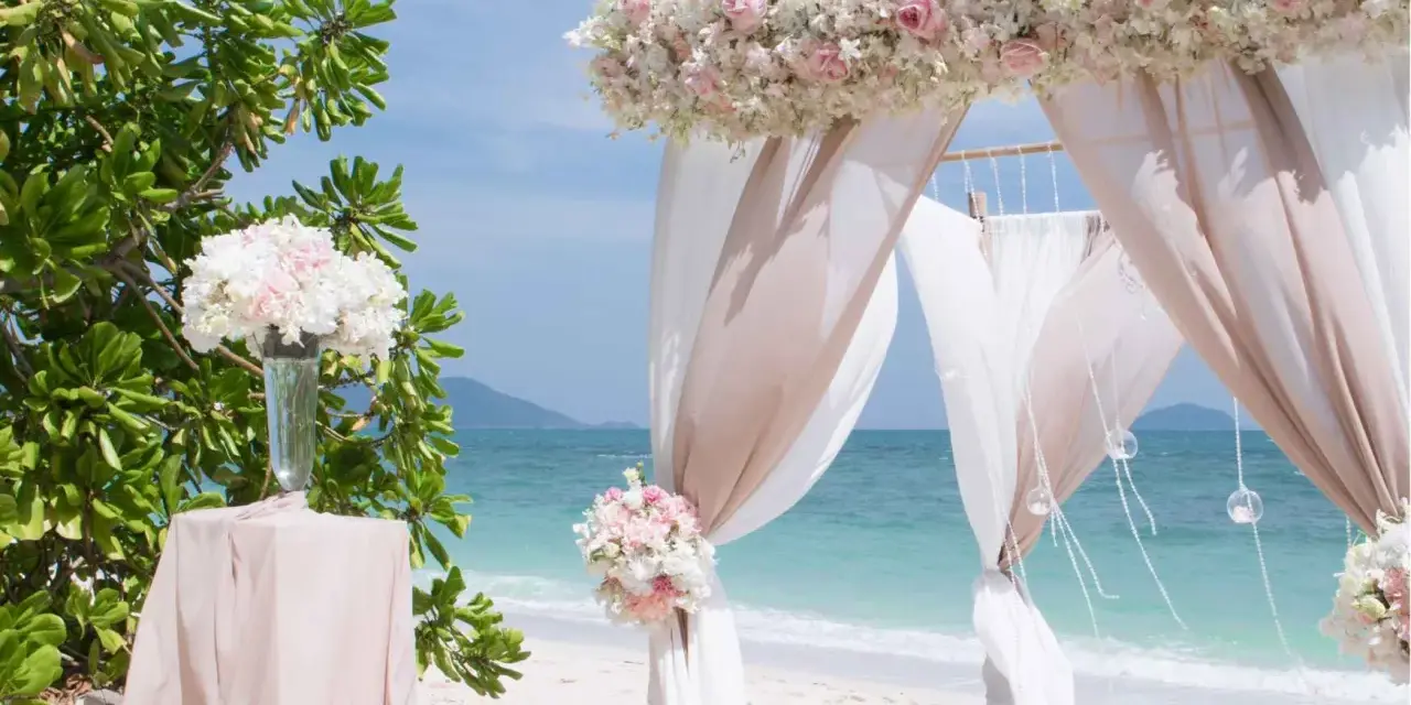 Romantyczny ślub na plaży. Biały baldachim ozdobiony różowymi i białymi kwiatami, zwiewne tkaniny i bukiety tworzą magiczną scenerię.