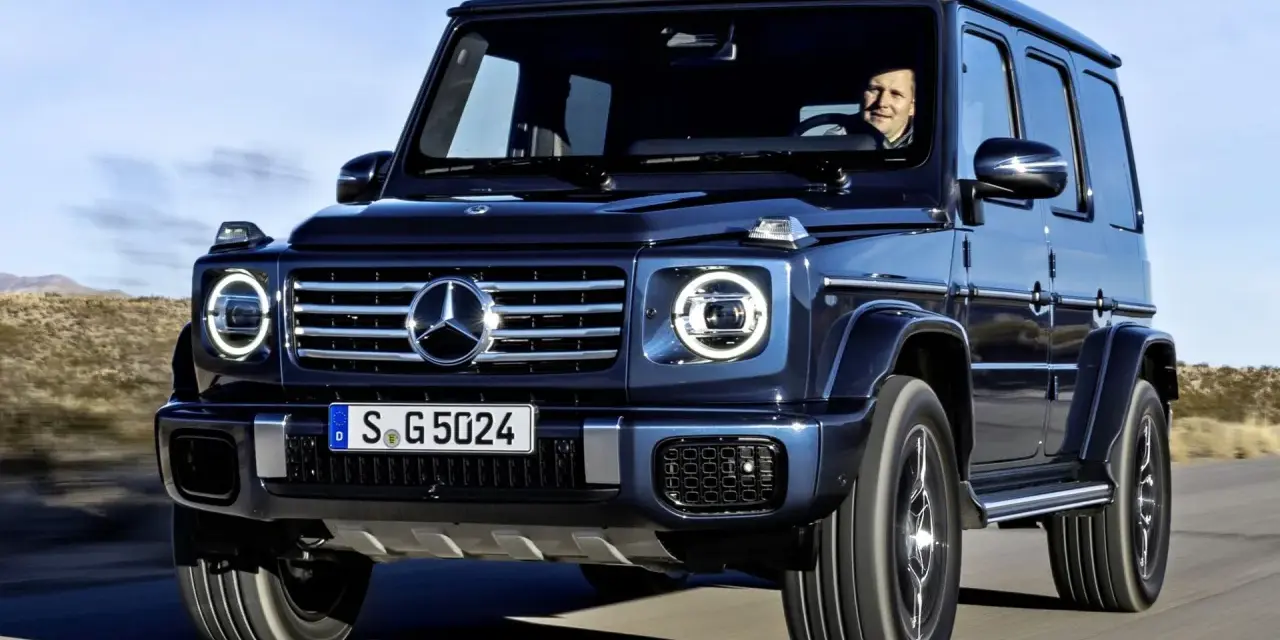 Ciemnoniebieski Mercedes G Klasa jedzie po pustynnej drodze. Kierowca uśmiecha się. Sprawdź mercedes g klasa cena!