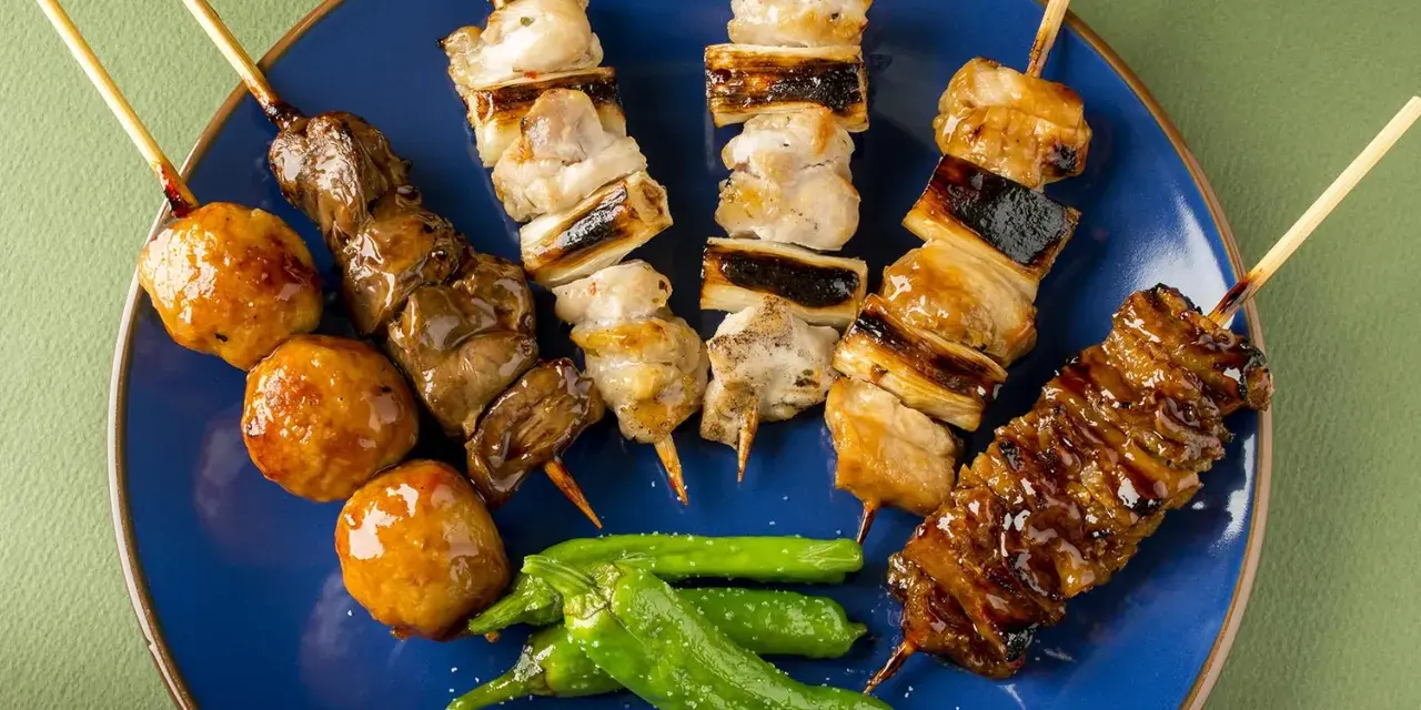 Różnorodne yakitori na niebieskim talerzu: kurczak, wątróbka, klopsiki i papryczki shishito.