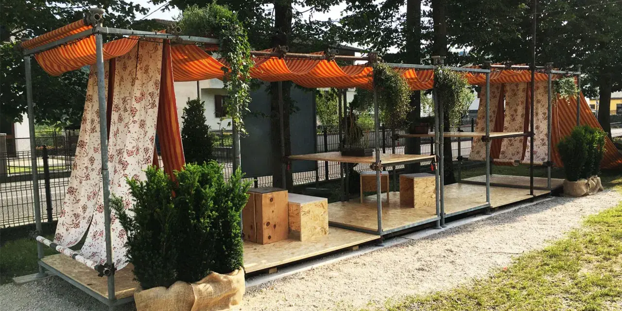 Struttura esterna con tende a righe e floreali, tavoli e sedute in legno. Un esempio di come costruire un gazebo con tubi innocenti, pratico e decorativo.