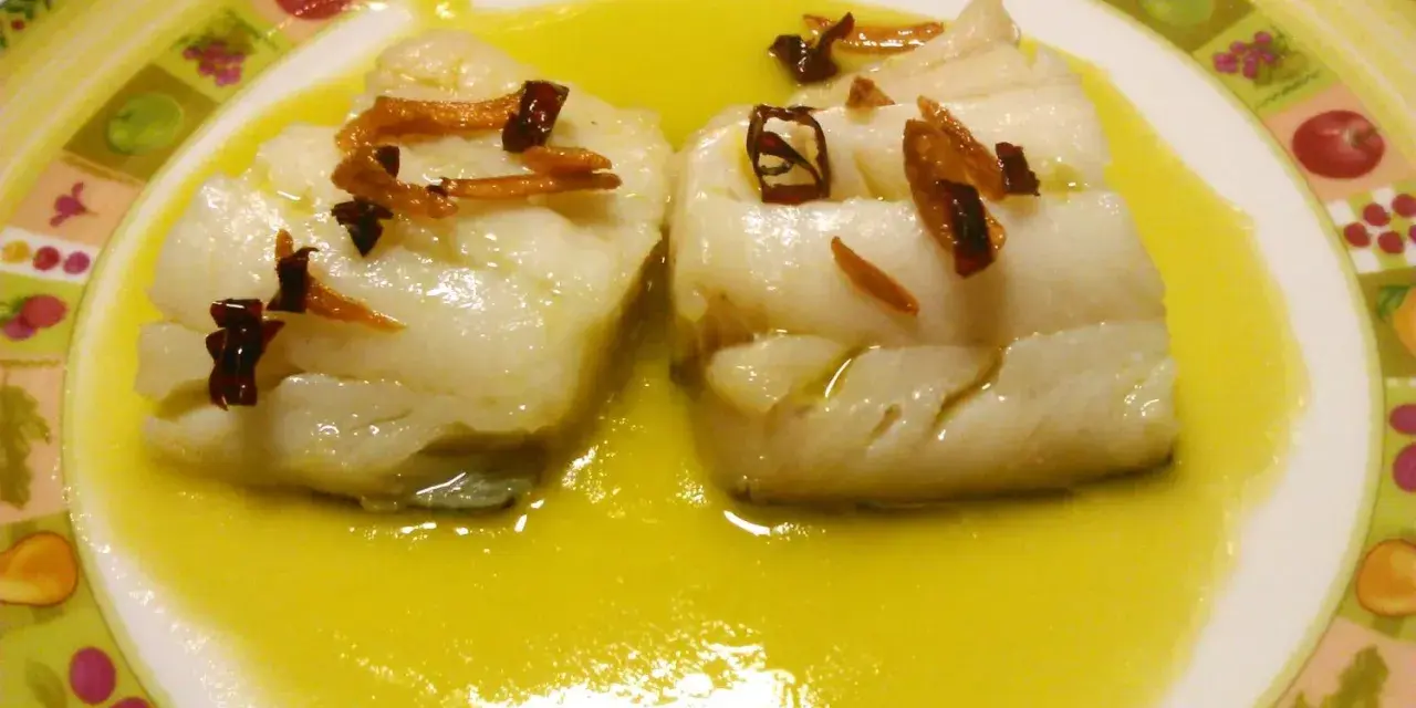 Dos trozos de bacalao al pil pil, cubiertos con ajo frito y guindilla, nadan en una salsa dorada sobre un plato decorado.