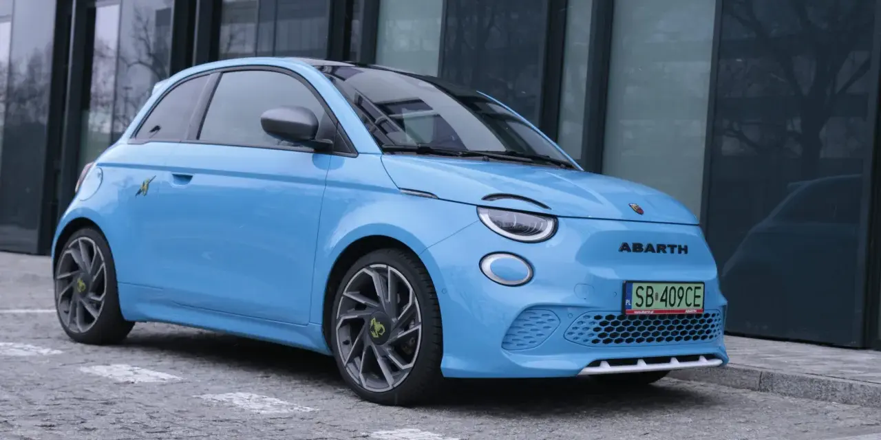 Niebieski Abarth 500e, gotowy do testów, zaparkowany przed nowoczesnym budynkiem.