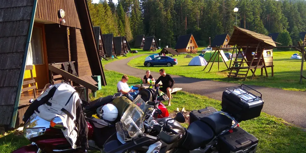 Motocykliści odpoczywają na kempingu w Rumunii, otoczeni drewnianymi domkami i lasem.