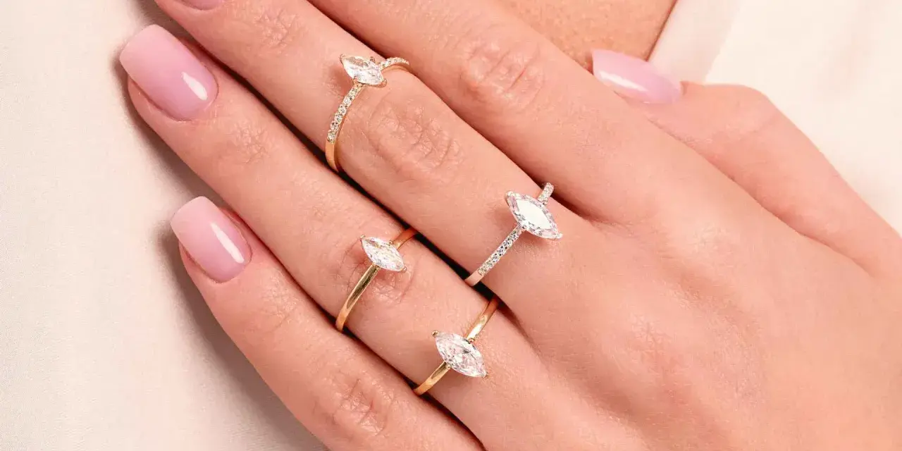 Dłonie z różowym manicure ozdobione są trzema złotymi pierścionkami z białymi kamieniami. Jeden z nich to pierścionek kwadratowy.
