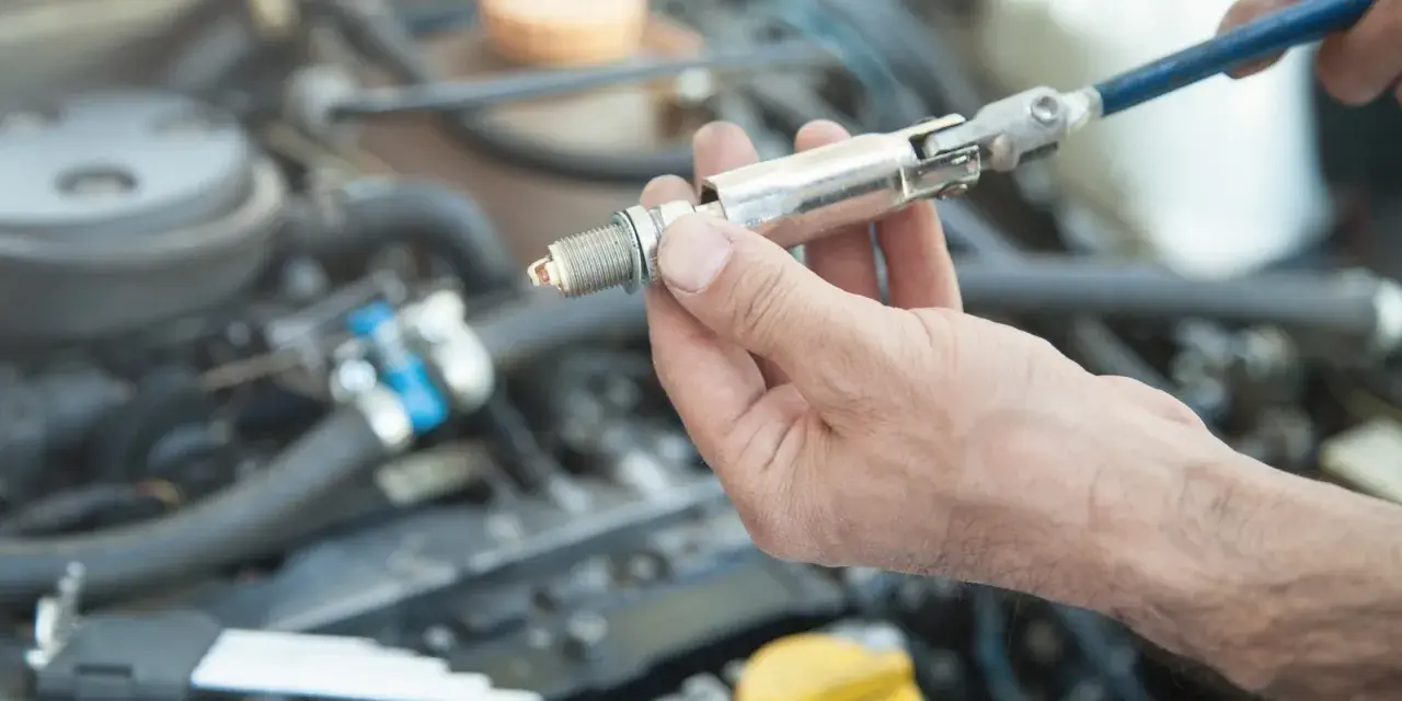 A hand holds a spark plug with a socket wrench attached, ready for a spark plug replacement. This DIY task can save on the spark plug replacement cost.