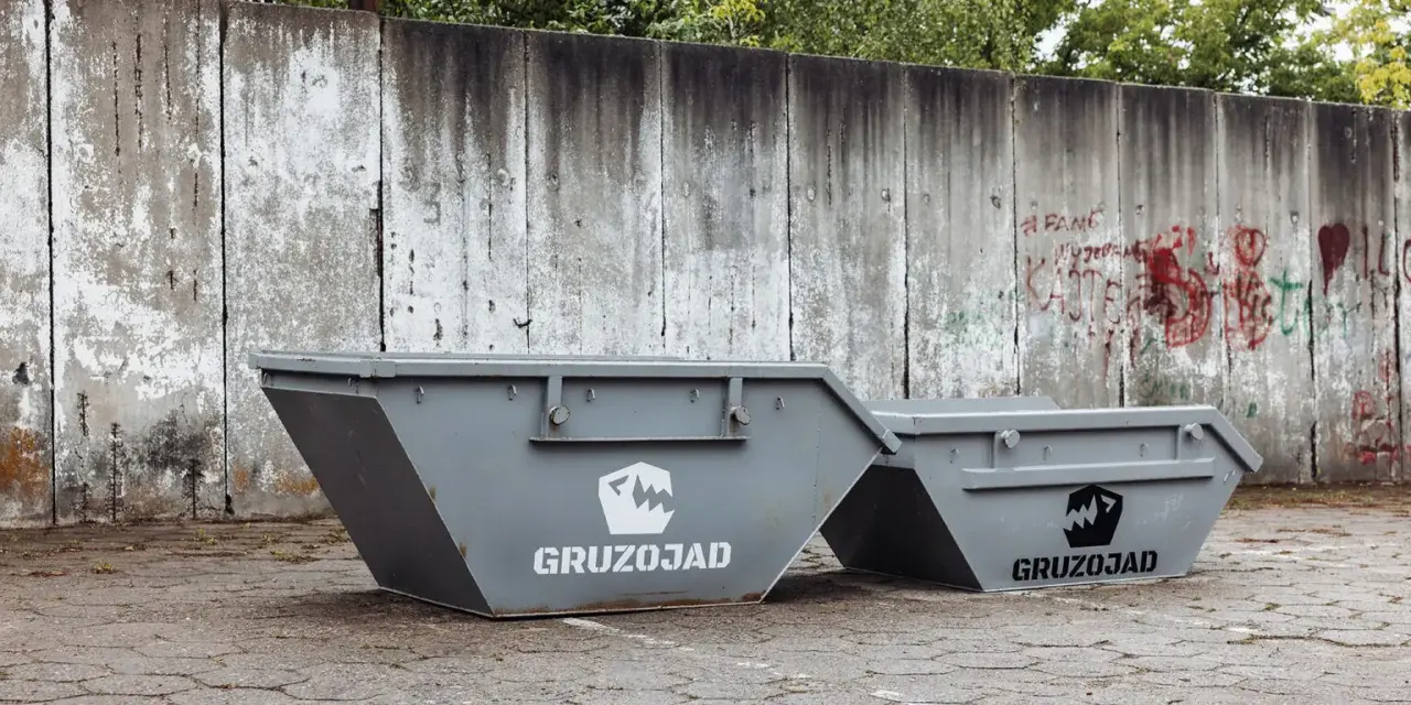 Dwa szare kontenery na śmieci zmieszane, z logo GRUZOJAD, stoją przed zniszczonym betonowym murem pokrytym graffiti.
