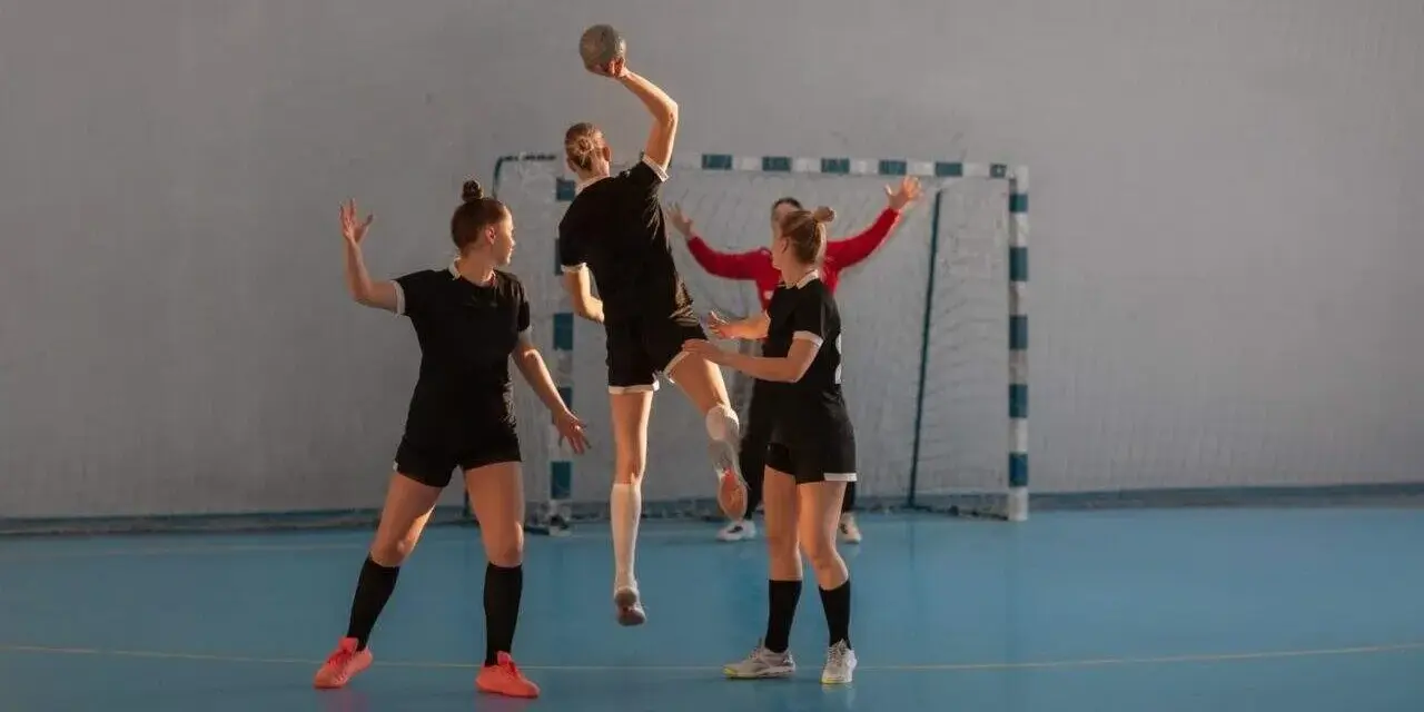 Piłkarki ręczne w akcji. Jedna z nich skacze do rzutu, podczas gdy koleżanki z drużyny i przeciwniczka obserwują. To dynamiczna gra pasywna w piłce ręcznej.