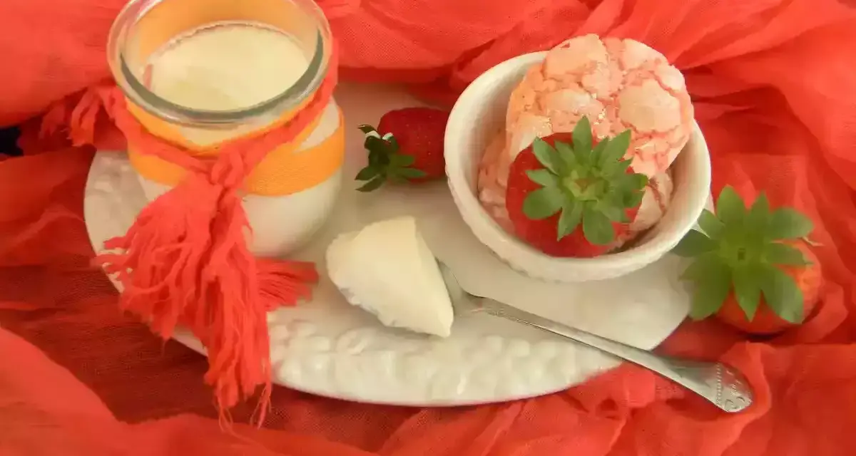 Yogur cremoso en frasco con fresas y trozos de yogur en un plato blanco sobre tela naranja.