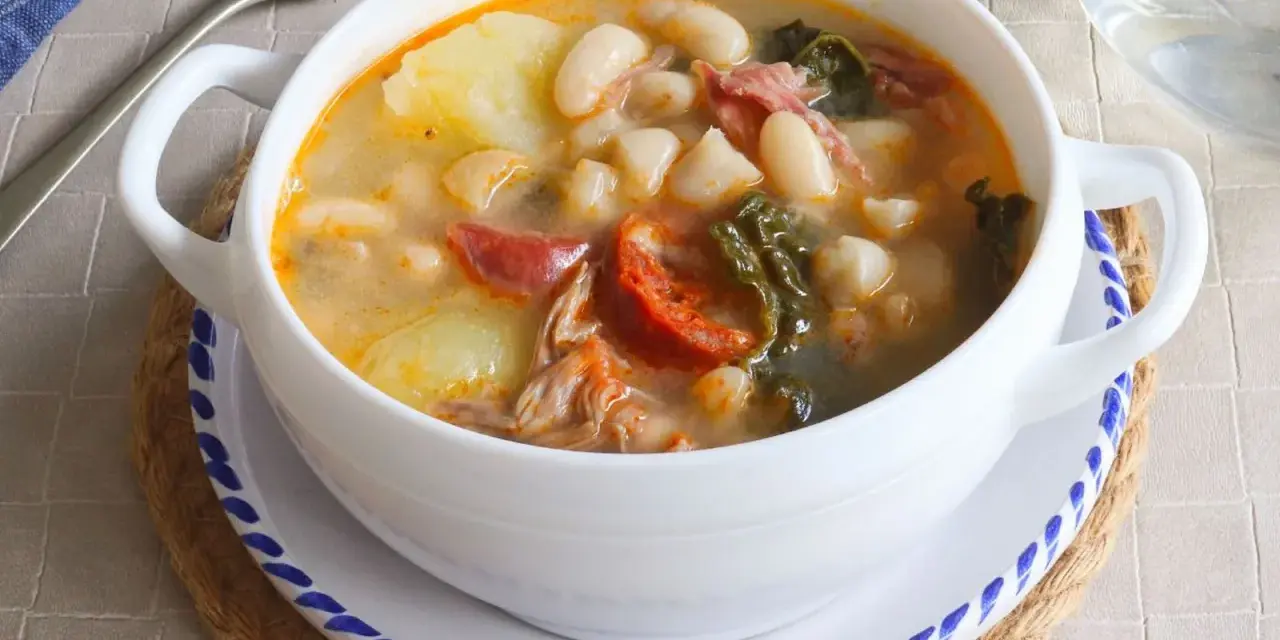 Un tazón blanco de caldo gallego con patatas, alubias, chorizo y grelos.
