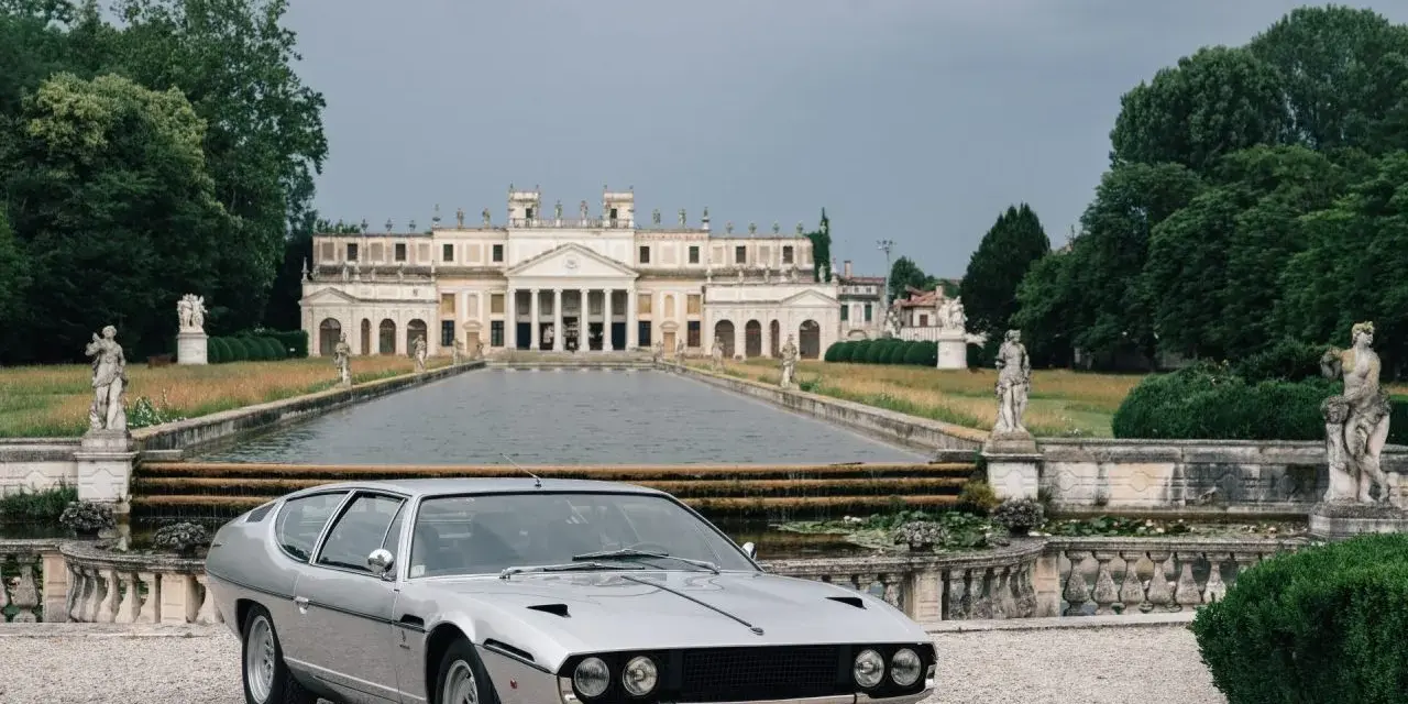 Srebrny Lamborghini Espada z lat 70. przed okazałą willą z basenem i rzeźbami.