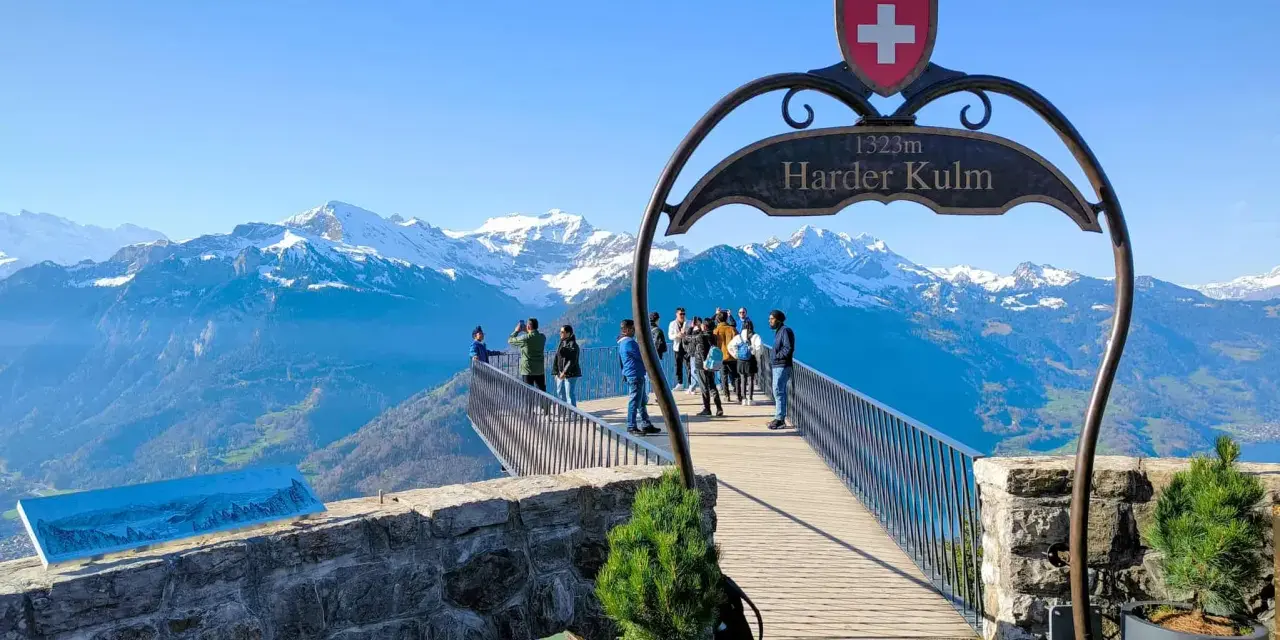 Tourists enjoy the breathtaking panorama from the Harder Kulm viewpoint, with snow-capped mountains under a clear blue sky.