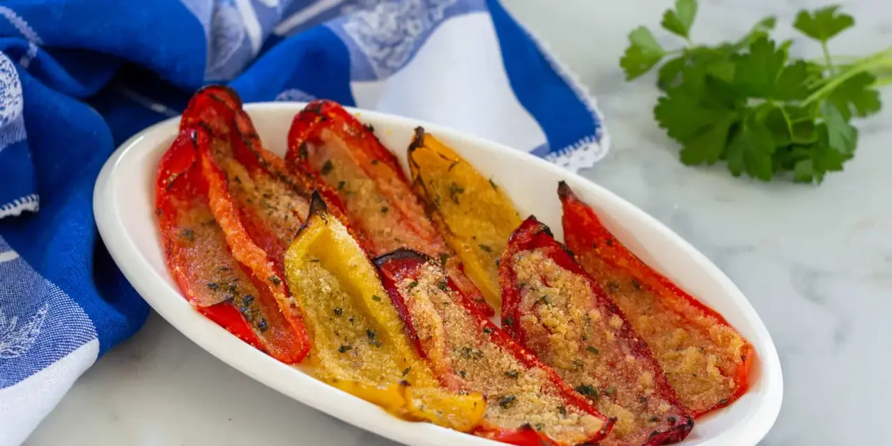 Peperoni gratinati al forno, rossi e gialli, cotti alla perfezione con pangrattato e erbe aromatiche, serviti su un piatto bianco con un tovagliolo blu.